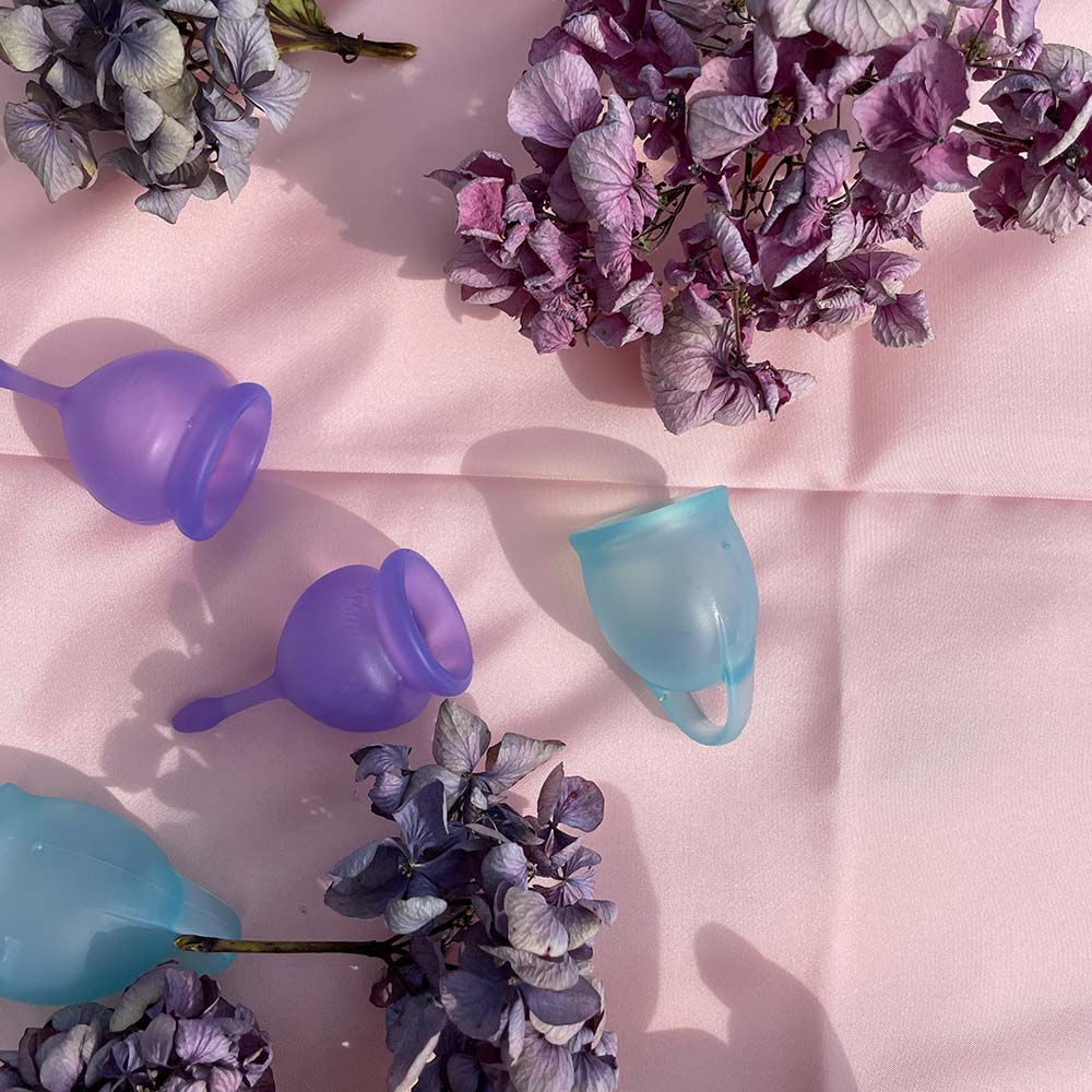 Feel good sæt med 2 menstruations kopper i violet og blå, på lyserødt tekstil med blomster